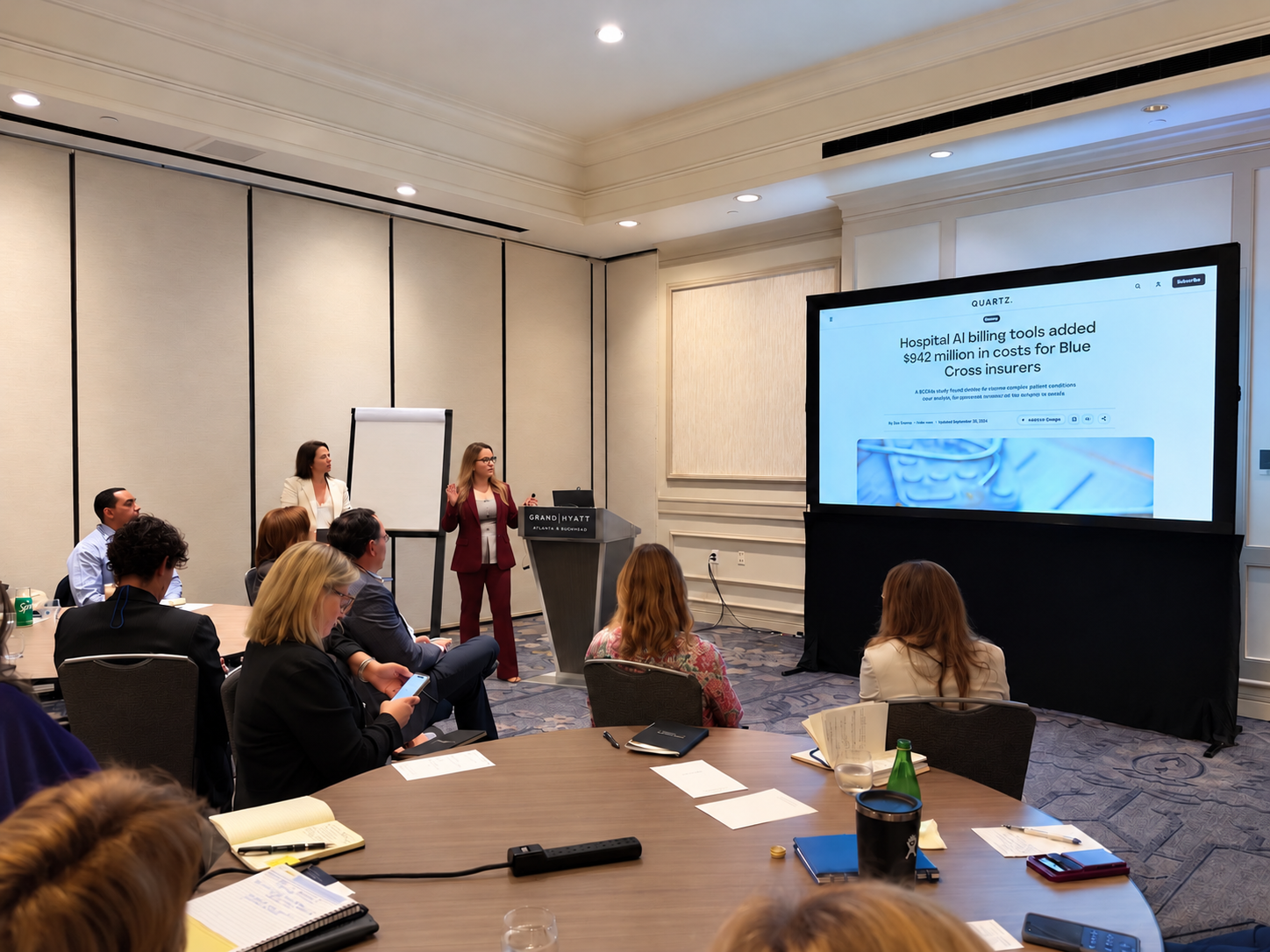 Two presenters lead a session in a hotel conference room. Directors sit at round tables with notebooks, and a large screen shows a Quartz headline about hospital AI billing tools adding $942 million in costs for Blue Cross insurers.