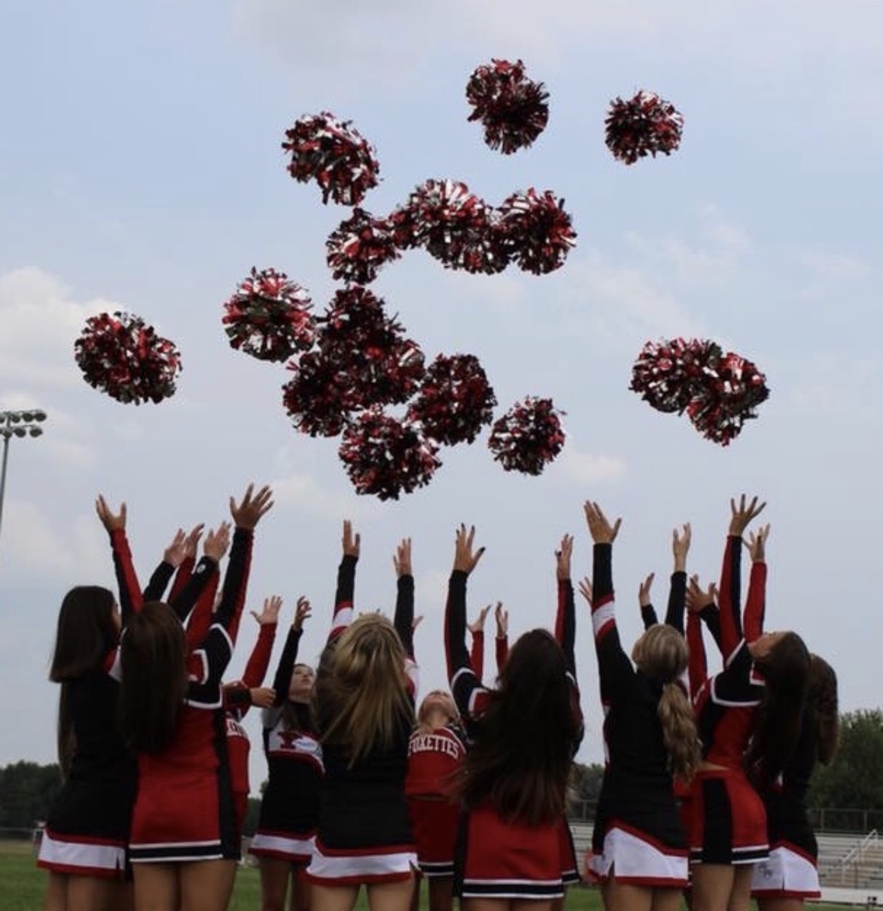 A high school dance team in red and black uniforms, seen from behind, throwing their pom-poms into the sky.